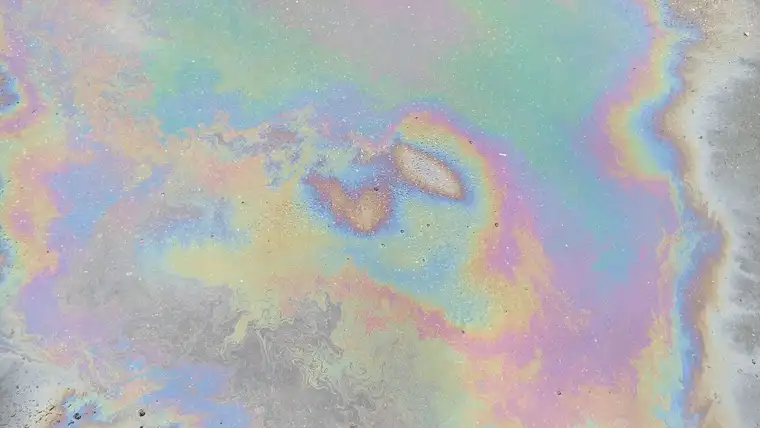 Rainbow sheen of leaked motor oil spreading across wet gray pavement