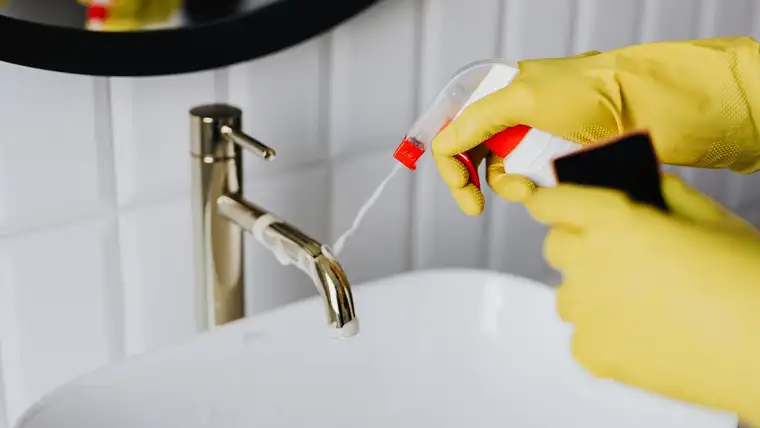 Yellow-gloved hands spraying cleaner onto a brass bathroom faucet above a white basin