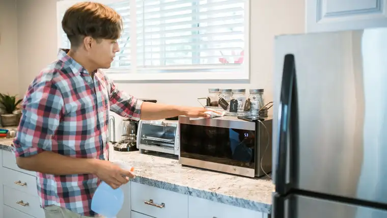 Man spraying and wiping a countertop microwave beside a toaster oven and fridge