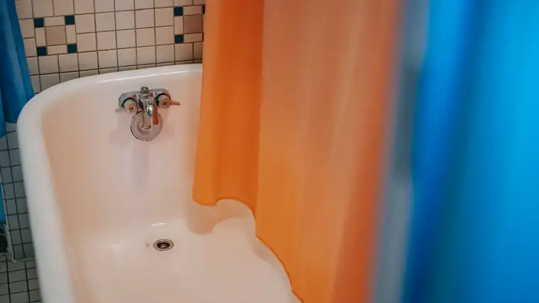 Old enamel bathtub with worn faucet and faint stains beside an orange shower curtain