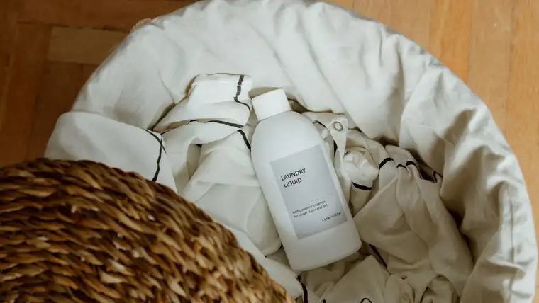 Bottle of liquid laundry detergent lying on white bedding in a woven hamper