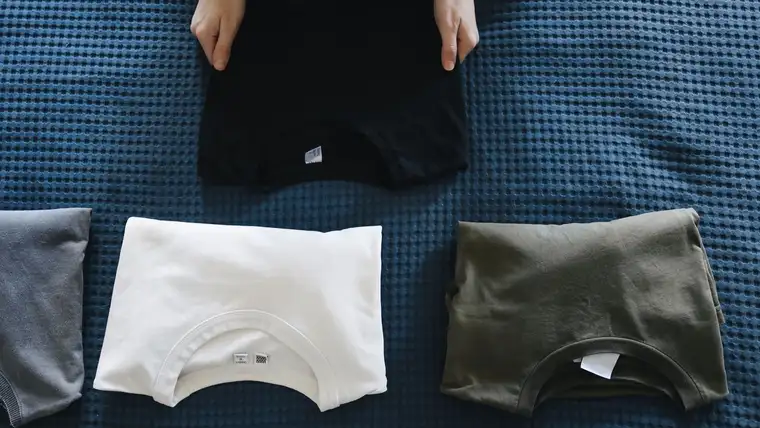 Hands folding black, white and olive cotton T-shirts on a blue bedspread