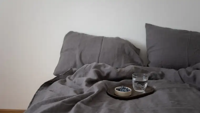 Rumpled gray linen bedding with a tray holding blueberries and a glass of water