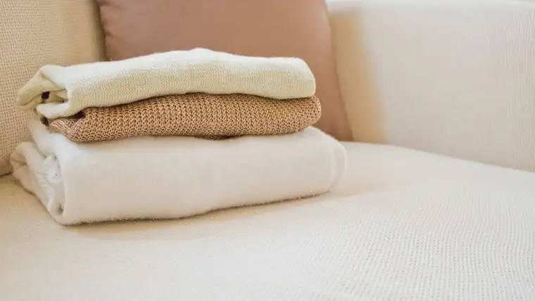 Folded cream and tan wool sweaters stacked on a light sofa cushion