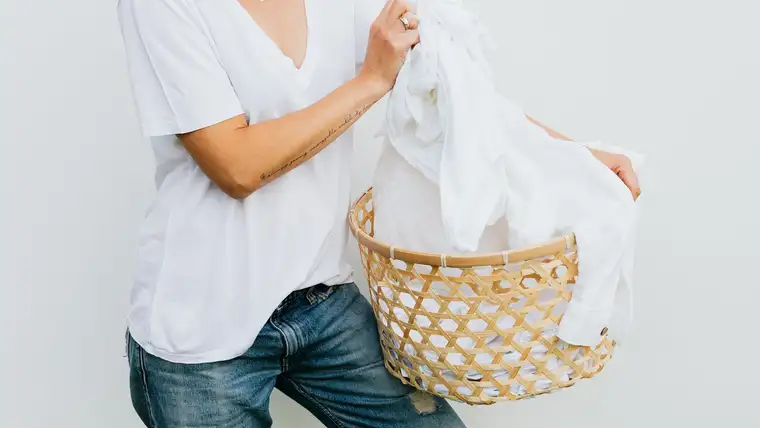 Woman carrying a woven basket piled with white laundry against a white wall