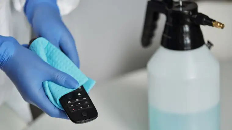 Gloved hands wiping a remote with a cloth beside a pump sprayer of cleaning solution