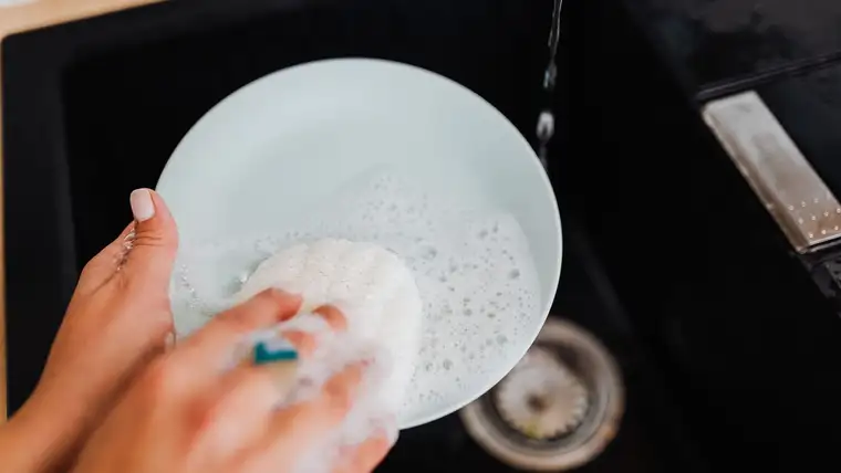 Hand washing a white plate with a sudsy sponge over a black sink