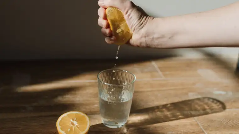 Hand squeezing a lemon half into a glass of water on a sunlit wooden table