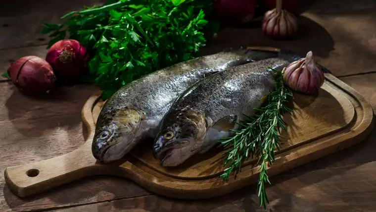 Two whole raw fish on a wooden cutting board with herbs, onions and garlic