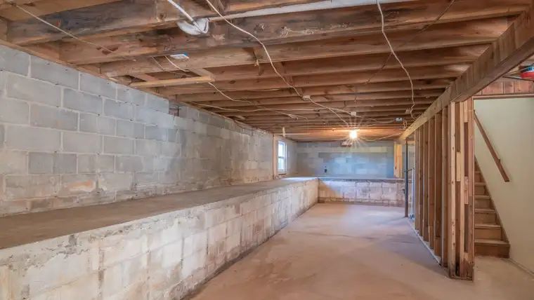 Empty unfinished basement with concrete block walls, wooden joists and stairs