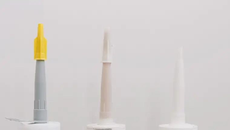 Nozzles of three caulk tubes in gray, beige and white against a white wall