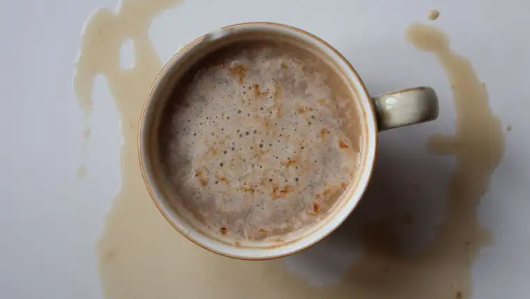 Cup of milky coffee sitting in a puddle of spilled coffee on white