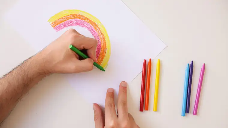 Hand coloring a rainbow with wax crayons on white paper, more crayons beside