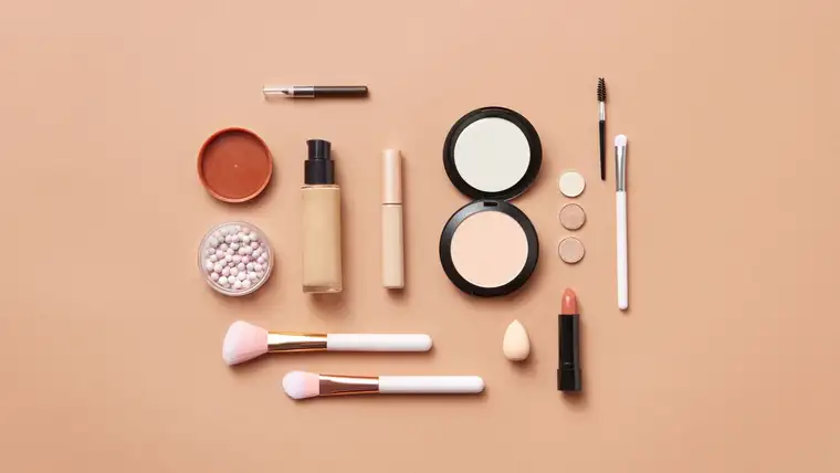 Foundation bottle, powder compact, lipstick and makeup brushes laid out on peach