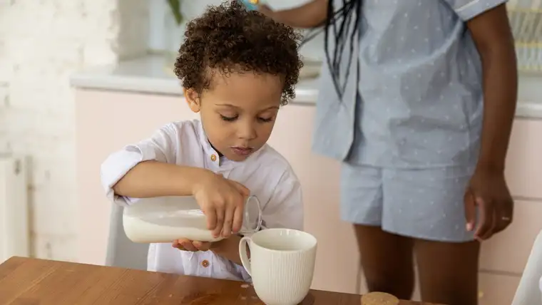 Young boy pouring milk into a mug, with milk spilled on the wooden table