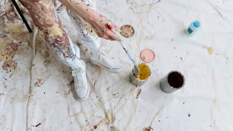 Paint-splattered legs and sneakers beside open paint cans on a dripping drop cloth