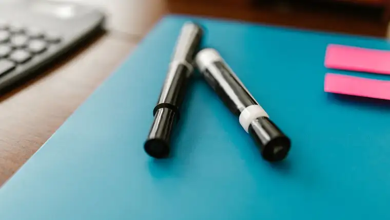 Two black permanent markers lying on a teal folder beside pink sticky notes