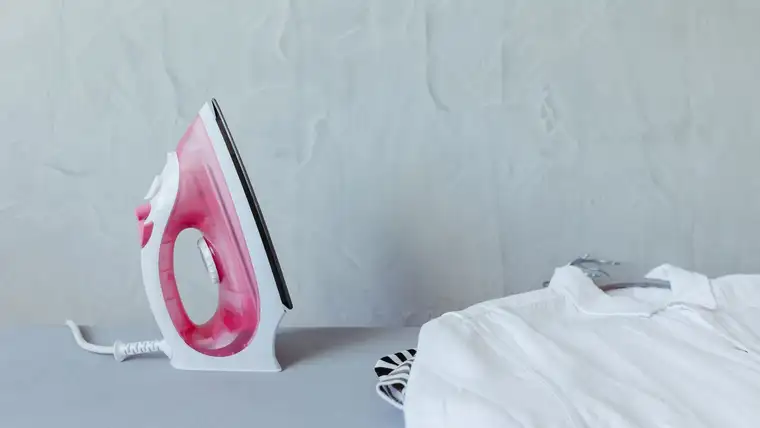 Pink steam iron standing on an ironing board next to a white shirt