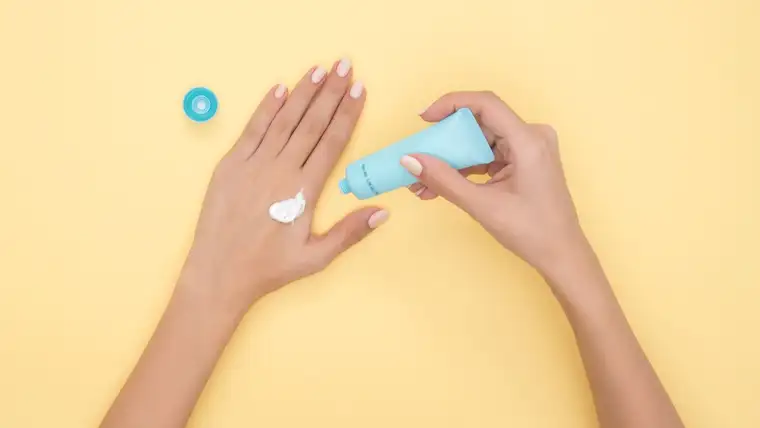 Hands squeezing white lotion from a blue tube onto the back of a hand