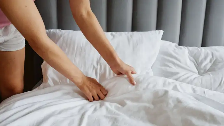 Hands smoothing crisp white sheets and pillows on a bed with gray headboard